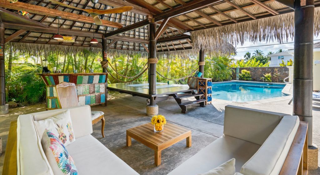 Tropical outdoor patio with thatched roof, white sofas, ping pong table, hammocks, and a pool surrounded by lush greenery in Pasco County